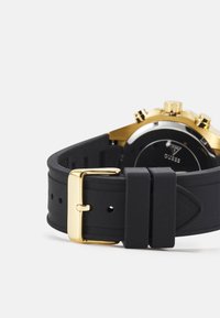 Cinturino in gomma nera con fibbia in metallo dorato, caratterizzato da una forma curva e dettagli testurizzati, che completa l'hardware dorato dell'orologio.