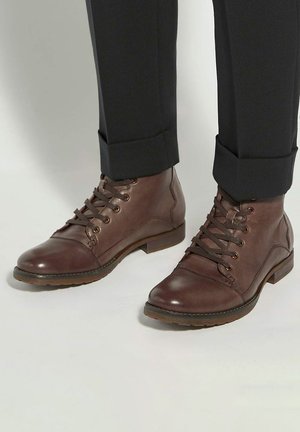 Brown leather ankle boots with round toes, lace-up front, and contrasting stitching, paired with dark rolled-up trousers.