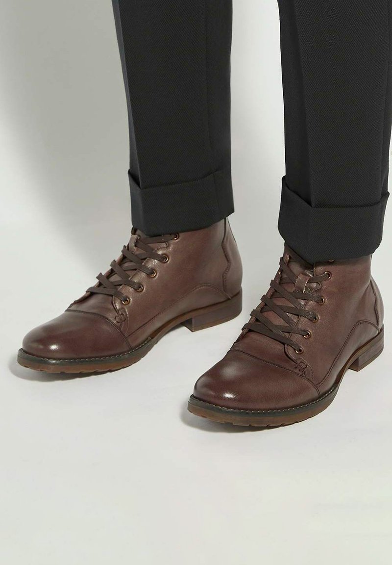 Brown leather ankle boots with round toes, lace-up front, and contrasting stitching, paired with dark rolled-up trousers.