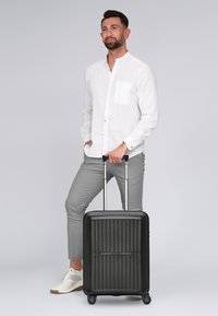 Maleta rígida negra con ranuras verticales, asa extensible y cuatro ruedas. Posicionada junto a un hombre con una camisa blanca y pantalones grises.