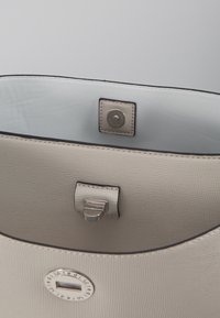 Sac à main beige clair avec une surface texturée. Il possède une doublure intérieure blanche, des accessoires en métal argenté et une fermeture à pression avec un accent de logo rond.