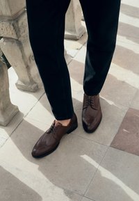 Brązowe skórzane buty formalne z detalami brogue, w połączeniu z czarnymi spodniami, na tle wzorzystej kamiennej podłogi.