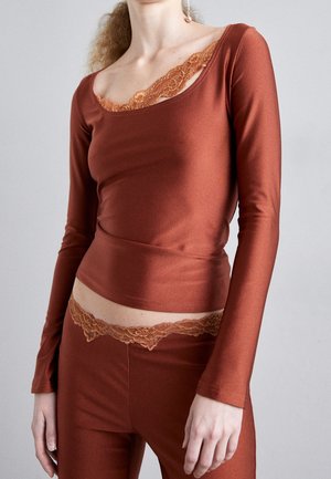 Kobieta ubrana w dopasowaną, rdzawego koloru bluzkę z długim rękawem i spodnie z dopasowaną koronkową lamówką na dekolcie i w pasie, stojąca na szarym tle.