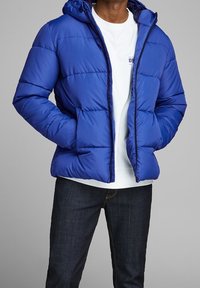 Niebieska pikowana kurtka puchowa z kapturem, z przodu zapinana na zamek. Nosi się ją na białej koszuli i łączy z ciemnymi dżinsami.