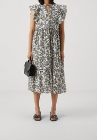 Midi šaty s květinovým potiskem v černobílé barvě, s volánovými rukávy, výstřihem ve tvaru srdce a nařaseným pasem; kombinované s černou kabelkou a sandály.