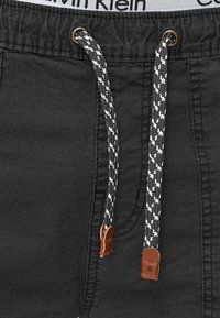 Shorts noirs en tissu texturé, dotés de cordons de serrage en corde blancs et noirs contrastants, avec des accents en cuir aux extrémités.