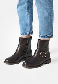 Bottines en cuir marron avec une fermeture éclair sur le côté, semelle plate, et une sangle décorative avec une boucle en or. Portées avec un jean bleu retroussé.