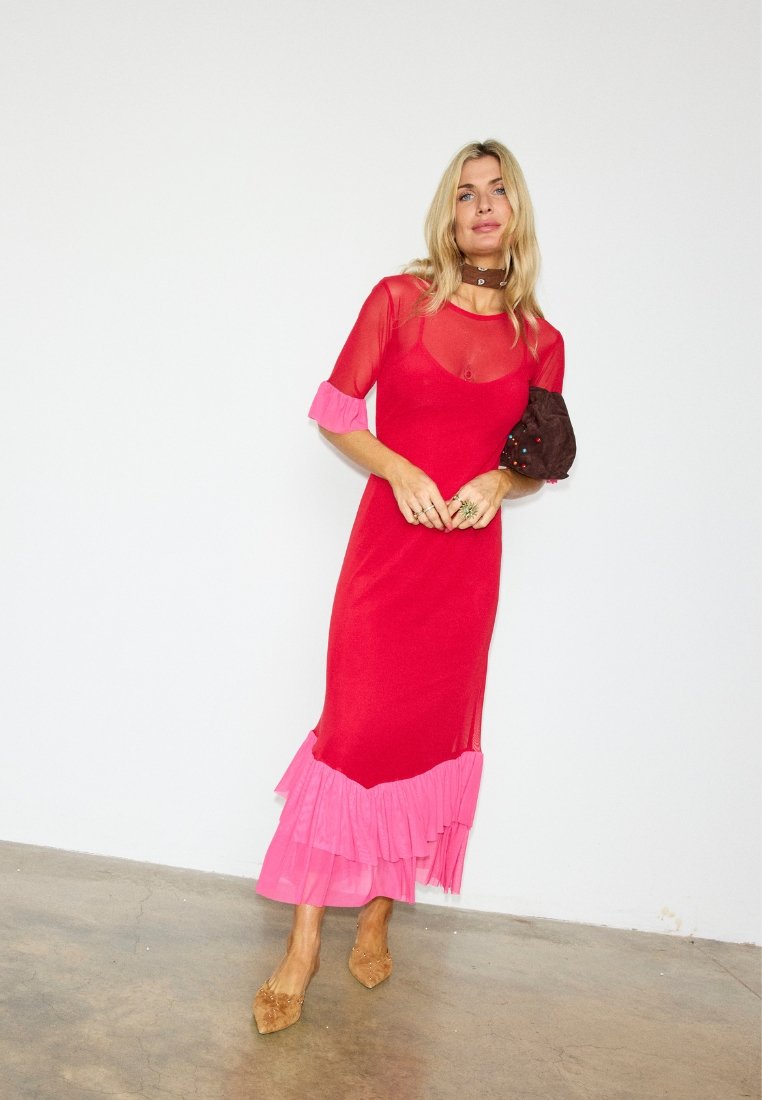 Femme aux cheveux blonds portant une robe rouge avec des volants roses, manches transparentes, un collier brun, tenant une pochette brune cloutée, debout sur un sol en béton.