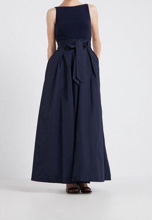 Kvinde iført ærmeløs marineblå jumpsuit med vide ben og bundet taljebælte samt sorte åbne hæle, stående mod en hvid baggrund.