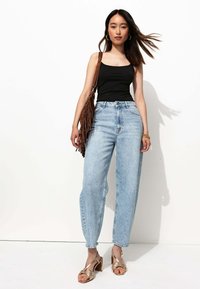 Jean taille haute bleu clair, débardeur noir ajusté, sandales à talons dorées à lanières et sac à épaule en franges marron sur fond blanc.