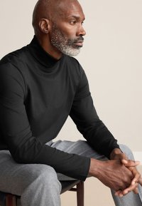 Sort langærmet rullekraveskjorte med glat tekstur, kombineret med lysegrå bukser, siddende på en træstol mod en neutral baggrund.