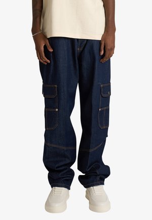 Persona che indossa pantaloni cargo in denim blu scuro con molte tasche e sneakers bianche, in piedi davanti a uno sfondo neutro.