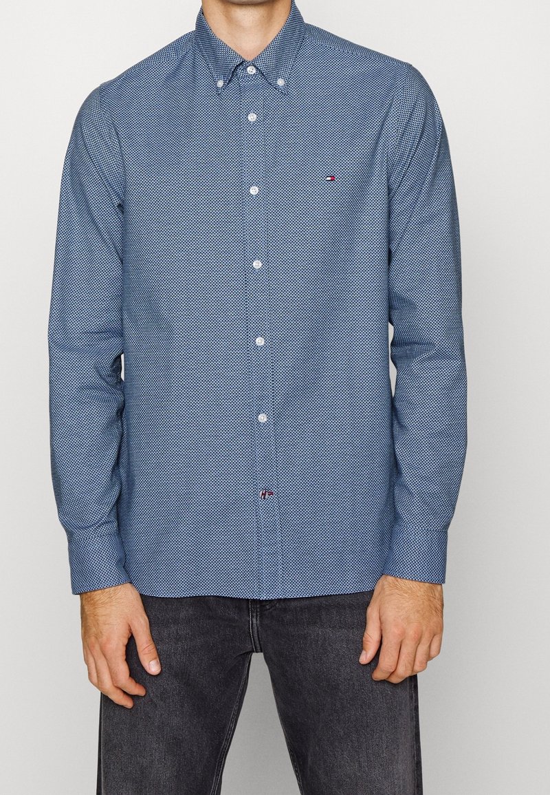 Homme portant une chemise bleue à manches longues à motif avec un petit logo sur la poitrine, associée à un jean gris foncé, debout devant un fond uni.
