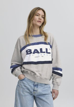 Sivi dukserica s mornarskoplavim i bijelim prugama, s velikim "BALL" logotipom preko prsa. Uparena s svijetloplavim trapericama.