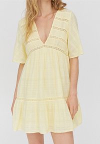 Gelbes kurzärmliges Kleid mit tiefem V-Ausschnitt, strukturiertem Stoff und Spitzenakzenten unter der Brust sowie am Saum. Ausgestellte Silhouette.