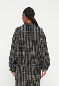 Svart rutig jacka med en texturerad tyg, som har en krage, sänkta axlar och elastisk midja. Långa ärmar med samlade manschetter.