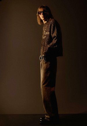 Hnedý pletený sveter s textúrovaným vzorom a logom, skombinovaný s voľnými hnedými nohavicami s dekoratívnymi akcentmi. Dokončené tmavými topánkami.