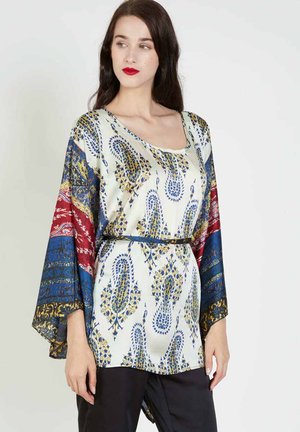 Mujer con cabello largo y oscuro lleva una túnica estampada con mangas anchas en tonos azules, rojos y dorados, combinada con pantalones negros sobre un fondo liso.