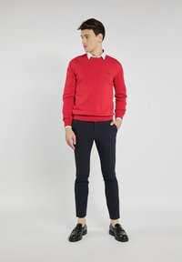 Czerwona sweter z prążkowanymi mankietami, biała koszula z kołnierzykiem pod spodem, granatowe slim-fit spodnie i czarne loafersy. Sweter posiada małe logo.
