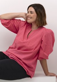 Blusa rosa con maniche a sbuffo e scollatura a V, caratterizzata da motivi floreali testurizzati su tutta la superficie. Abbinata a pantaloni scuri.