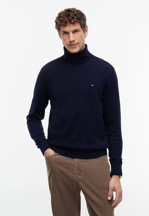 Homme aux cheveux bouclés portant un pull à col roulé bleu marine et un pantalon marron clair, debout avec une main dans la poche sur fond blanc.
