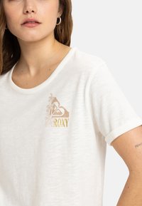 Camiseta blanca de manga corta con cuello redondo, que presenta un estampado de logo floral en dorado y marrón claro en la parte izquierda del pecho.