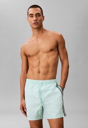 Sportlicher Mann mit kurzen Haaren, der hellblaue Badeshorts mit schwarzen und weißen Seitenstreifen trägt, steht vor einem schlichten grauen Hintergrund.