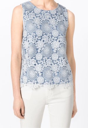 Niebieska bluzka bez rękawów z białą koronką w kwiatowy wzór, noszona z białymi spodniami, ukazująca górną część tułowia i ramiona na jednolitym tle.