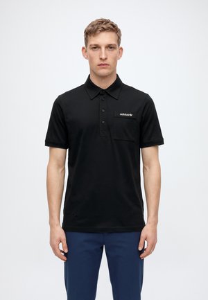 Un homme pose avec un polo noir à manches courtes Adidas, doté d’une poche poitrine, et un pantalon bleu marine, sur un fond uni clair.
