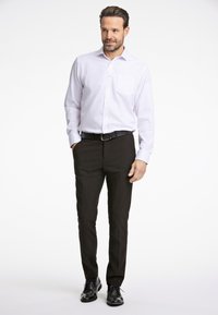 Wit overhemd met een borstzak, gecombineerd met zwarte broek en zwarte nette schoenen, staand op een effen achtergrond.