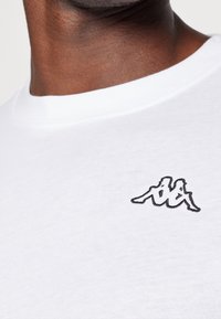 Bílý bavlněný tričko s černě vyšitým logem na levé hrudi. Tkanina je hladká a má kulatý výstřih.