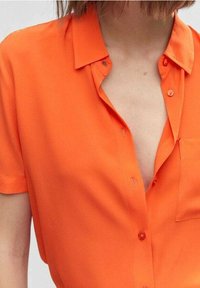 Pomarańczowa bluzka z krótkim rękawem, z kołnierzykiem, z zapięciem na guziki z przodu, kieszenią po lewej stronie oraz gładką teksturą materiału.