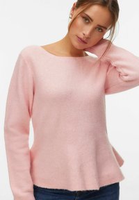 Jeune femme aux cheveux châtain clair portant un pull rose doux et un jean bleu, posant avec une main derrière la nuque.