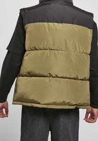 Pufferväst i svart och olivgrönt, med hög krage, kviltat mönster och glänsande, slät tyg. Bakvyn visar horisontella sömmar.