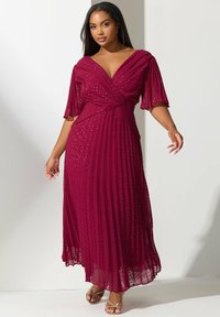 Mujer vistiendo un vestido largo hasta el suelo, plisado, de un magenta profundo con sutiles lunares y mangas transparentes hasta el codo, posando frente a un fondo blanco.