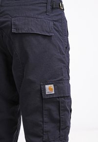 Granatowe spodnie cargo Carhartt z bocznymi kieszeniami na klapach i widocznym logo marki na dolnej kieszeni.