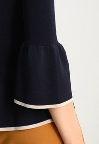 Granatowy dziany top z rozszerzanymi rękawami. Charakteryzuje się kremowym obszyciem na mankietach i dole, prezentując gładką fakturę.