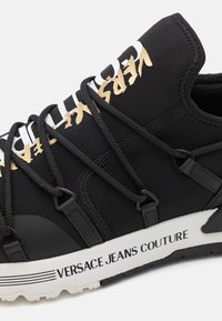 Melna sporta kedu ar elastīgām auklām, gumijas akcentiem un baltu starpzoli, kurā iespiests "VERSACE JEANS COUTURE". Gluda tekstūra.