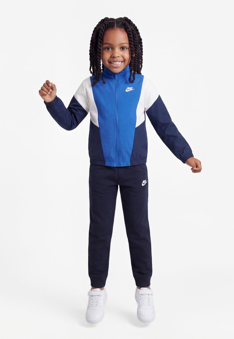 Lächendes Kind mit geflochtenen Haaren, das einen blau-weißen Nike-Trainingsanzug und weiße Turnschuhe trägt, steht mit leicht erhobenen Armen.