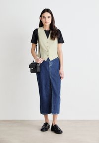 Gilet beige avec des boutons sur un t-shirt noir ; jupe en denim à mi-mollet avec une fente à l'avant ; mocassins noirs ; petit sac en bandoulière noir.