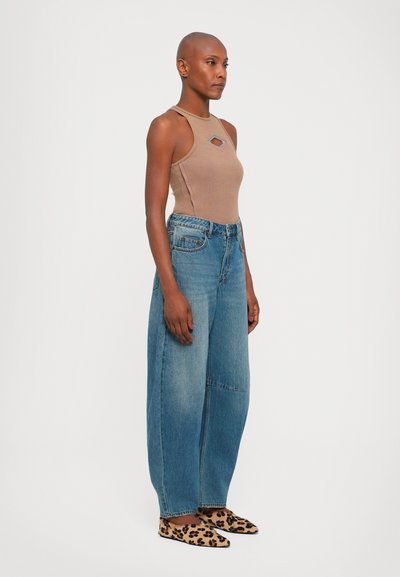 Tan halterneck bodysuit med diamantutskärning, matchad med lösa blå denimjeans. Platta skor med leopardmönster. Modellen står sidledes.