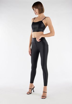 Nainen pukeutuneena mustaan keinonahkaseen lyhytpaitaiseen toppiin, jossa on ohuet olkaimet, korkeavyötäröisiin leggingsseihin ja mustiin korkokenkiin, seisoo valkoista taustaa vasten.