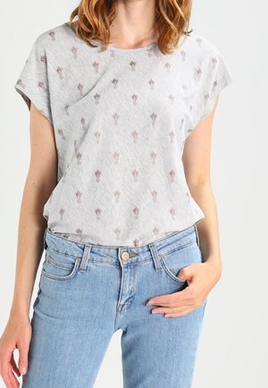 Kobieta w jasnoszarej koszulce z drobnymi brązowymi liściastymi wzorami, włożonej do jasnoniebieskich dżinsów, z ręką opartą na biodrze.