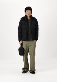 Schwarze gepolsterte Jacke mit Reißverschluss vorne, Kapuze und strukturierter Oberfläche, über einem dunklen Hemd getragen, kombiniert mit lockeren olivgrünen Hosen und einer schwarzen Tasche.