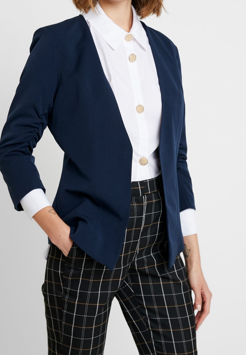 Femme portant un blazer bleu marine, une chemise blanche avec des boutons dorés et un pantalon noir à carreaux, les mains dans les poches, debout devant un fond blanc.