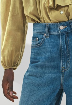Aukšto juosmens mėlyni džinsai iš denim, turintys penkias kišenes, metalinį mygtuką ir kontrastingą siuvimą, derinami su šviesiai alyvuogių spalvos bluzonu.