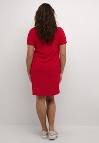 Robe en coton rouge avec un design simple, à manches courtes et longueur genoux. Portée avec des baskets beiges, montrant une texture lisse.