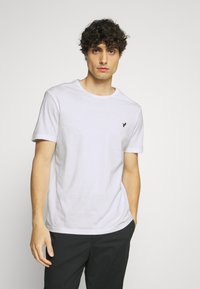 White short-sleeve t-shirt in soft cotton with a round neckline and a small black logo on the left chest. Model wears dark pants.