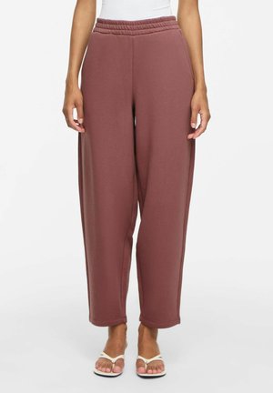 Femme portant un pantalon large, ample, taille haute, mauve avec une taille élastique et des sandales blanches sur un fond uni.