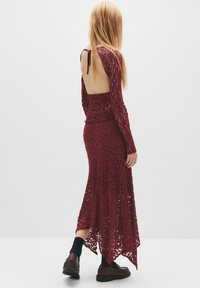 Bordeaux Spitzenkleid mit langen Ärmeln, einem tiefen Rückenausschnitt und einem High-Low-Saum. Getragen mit schwarzen Socken und dunklen Schuhen. Strukturierter Stoffdetails.
